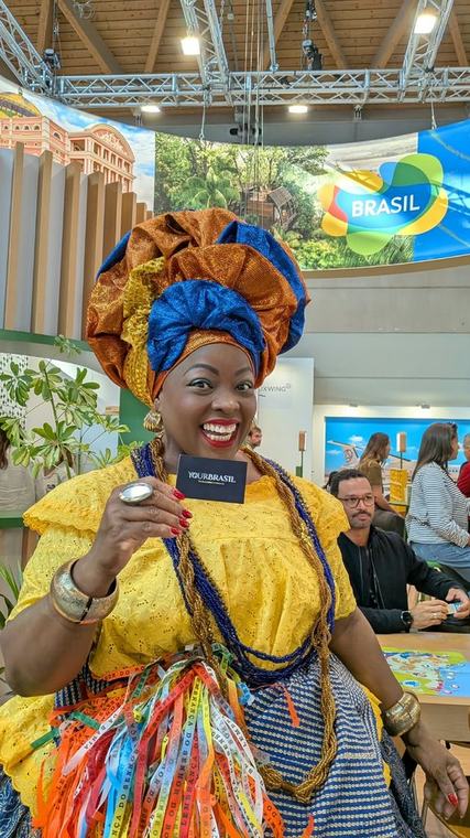 Woman in traditional Bahian dress holding a YourBrasil business card at the Brasil pavilion in Rimini